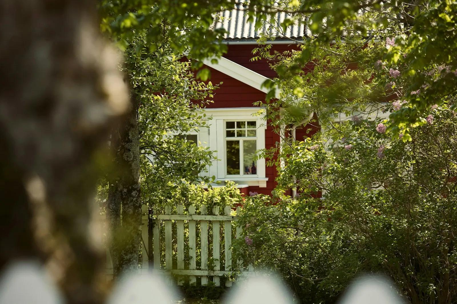 Rødt trehus bak et hvitt stakittgjerde og grønne trær